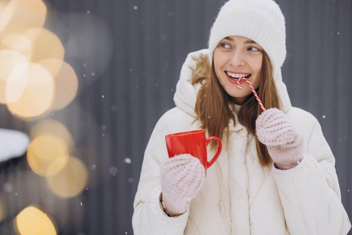 Happy woman in winter clothing holding hot cocoa and candy cane outdoors during holidays.