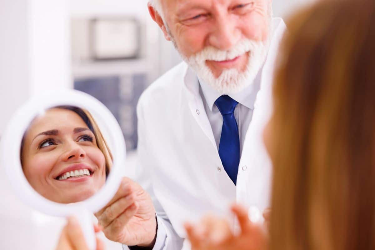 Elderly male dentist showing smiling female patient her reflection in handheld mirror after cosmetic dental treatment.