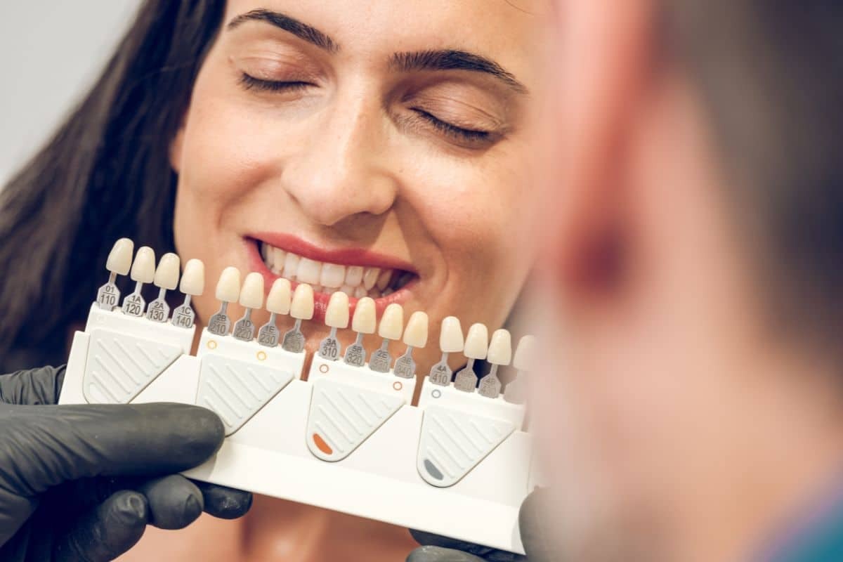 Woman examining dental veneer shade guide samples against her teeth for color matching.